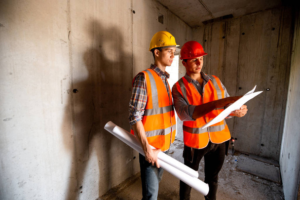 Stavbyvedoucí a inženýr oblečený v oranžové pracovní vesty a přilby práce s dokumentací stavby uvnitř budovy ve výstavbě - Fotografie, Obrázek