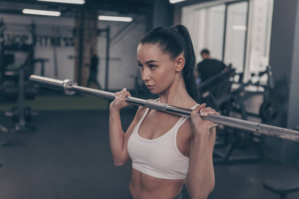Gorgeous fit and toned sportswoman lifting barbell at the gym, copy space on the side. Attractive female athlete in white sports bra exercising with barbell. Sexy sportswoman weightlifting - Zdjęcie, obraz