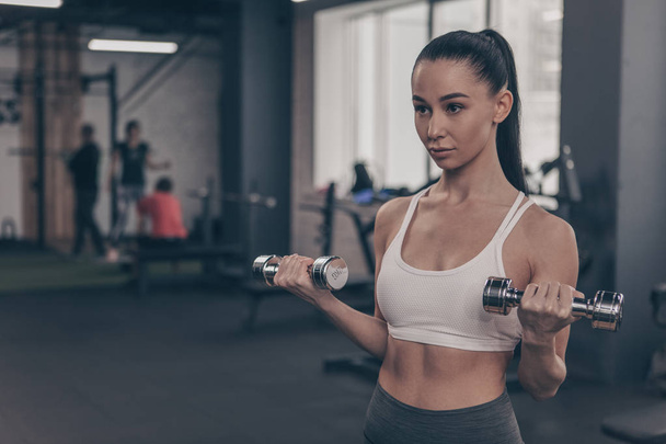 Attractive female athlete in sportswear exercising at the gym, lifting dumbbells. Beautiful fitness woman training at healthclub, copy space. Gorgeous athletic woman doing biceps exercises. Fitness concept - Photo, Image