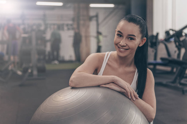 Ευτυχής όμορφη γυναίκα χαμογελά στη φωτογραφική μηχανή, αναπαύεται στο γυμναστήριο μετά από συνεργασία με μπάλα γυμναστικής. Χαρούμενα υγιή αθλήτρια χαλαρώνοντας στο σπορ στούντιο μετά την άσκηση, αντίγραφο χώρου - Φωτογραφία, εικόνα