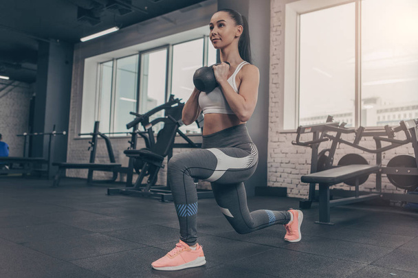 Low angle shot of a sporty young woman doing lunges with kettlebell. Attractive sportswoman enjoying working out at the gym, copy space. Fitness female in sportswear training with weights - Foto, Bild