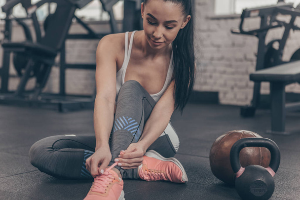 Cropped shot of a lovely young sportswoman tying her shoelaces, sitting on the gym floor, copy space. Beautiful fitness woman resting after working out at sports studio. Motivation, sportswear concept - Foto, Bild