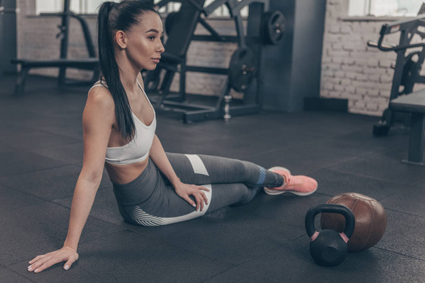 Full length shot of a gorgeous long haired sportswoman resting after exercising, sitting on gym floor, looking away thoughtfully. Beautiful fitness woman relaxing after workout, copy space. Health concept - Photo, Image