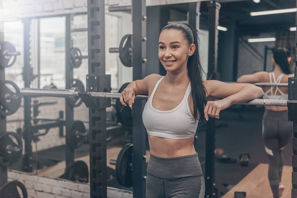 Die gesunde, fröhliche junge Fitnessfrau lehnt an einer Langhantel im Fitnessstudio, lächelt fröhlich, kopiert den Raum. schöne sexy Sportlerin entspannt sich nach dem Training - Foto, Bild