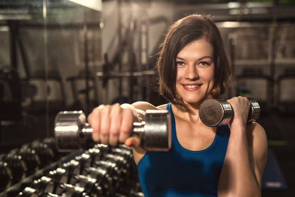 Happiness of hard work. Young beautiful happy woman training with dumbbells at the gym smiling to the camera joyfully selective focus copyspace vitality lifestyle sport people healthcare motivation  - Photo, Image