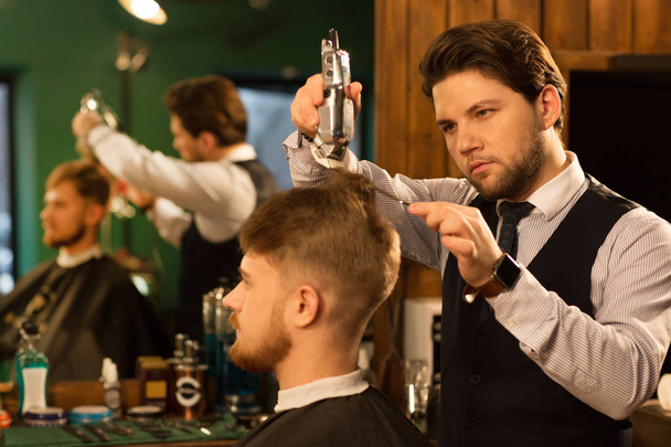 Attractive bearded professional barber giving his male client a haircut using electric trimmer clipper professionalism styling hairdressing occupation career job traditional barbering barbershop service. - Foto, Imagen