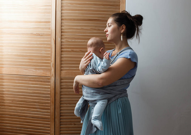 Młoda matka ubrana w lekki t-shirt niebieski i spódnica trzyma jej mały syn na ramionach w pokoju obok drzwi drewniane - Zdjęcie, obraz