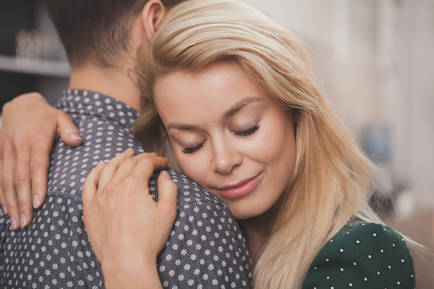 Close up of a beautiful woman smiling with eyes closed hugging her man tenderly. Charming happy woman embracing her loving husband. Couple cuddling at home. Love, romance, emotions concept - Photo, image