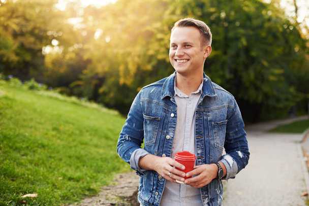 Young Hipster man standing with takeaway coffee in the park,  smiling plesantly into camera.  Happy carefree handsome guy in blue denim jacket - Foto, imagen