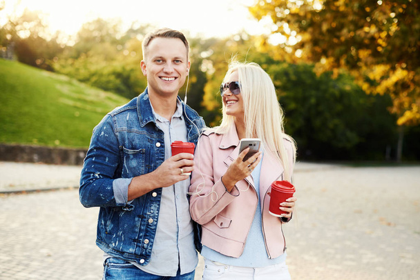 Smiling couple in love walking in autumn park, holding hands. Listen to music on headphones and drink coffee to go. Have a fun together. Hugging and kissing  - Foto, imagen