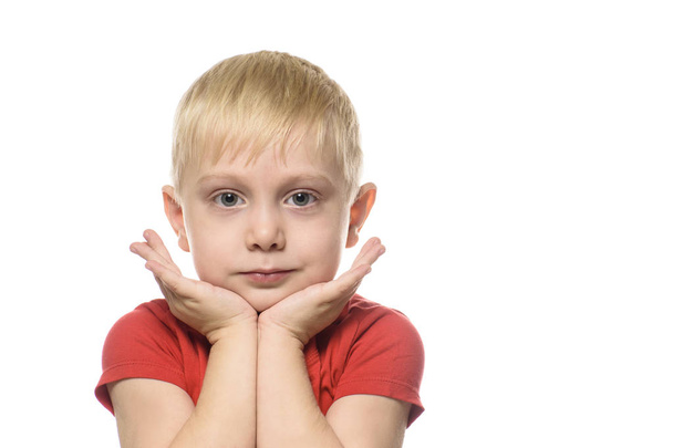 Συλλογισμένος ξανθό αγόρι σε ένα κόκκινο μπλουζάκι διπλωμένα τα χέρια του στο πηγούνι του. Απομονωθεί σε λευκό φόντο - Φωτογραφία, εικόνα