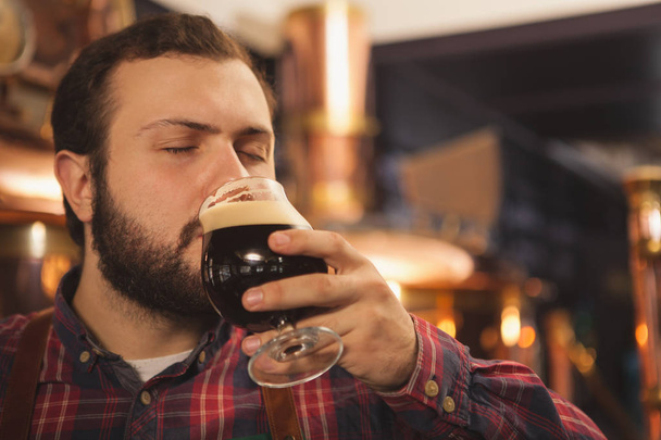 Close up van een brouwer nippen heerlijk bier met zijn ogen gesloten, kopie ruimte. Professionele beermaker genieten van werken bij zijn brouwerij donker bier proeverij. Productie, productie, verkoop concept - Foto, afbeelding
