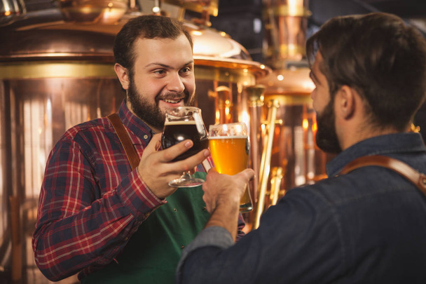 Бородатый пивовар разговаривает со своим помощником, работает на заводе по производству пива. Профессиональные пивовары звонят в пивные бокалы, празднуют успех. Малый бизнес, концепция предпринимательства
 - Фото, изображение