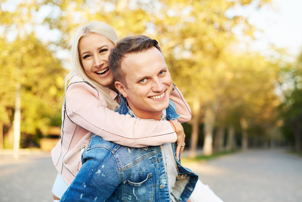 Happy couple on vacation. Lovers are laughing. Happy guy and girl. Lovers enjoy each other in the sunny park. Boyfriend carrying his girlfriend on piggyback - 写真・画像