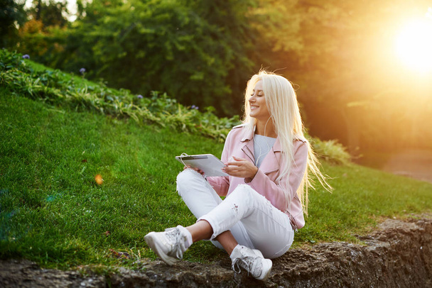 Smiling handsome woman listening relaxing music In the headphones  with her mobile phone or tablet at the park. A student listens to an online lecture and prepares for exams. girl enjoying loneliness - Fotoğraf, Görsel