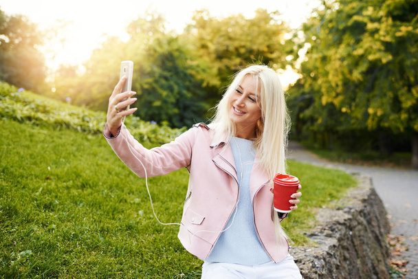 Ładny blond dziewczyna, słuchanie muzyki na słuchawki na smartfonie jest dobra zabawa i selfie w parku. Ona trzyma kawę iść.  - Zdjęcie, obraz