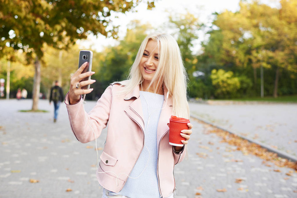 Sunny lifestyle fashion portrait of young stylish blond woman walking on street or park with phone, wearing trendy outfit, drinking coffee to go, smiling, enjoy weekends - Photo, Image