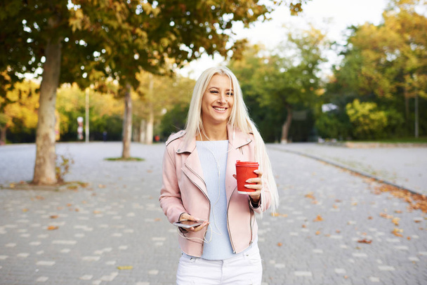 Sunny lifestyle fashion portrait of young stylish blond woman walking on street or park with phone, wearing trendy outfit, drinking coffee to go, smiling, enjoy weekends - Φωτογραφία, εικόνα