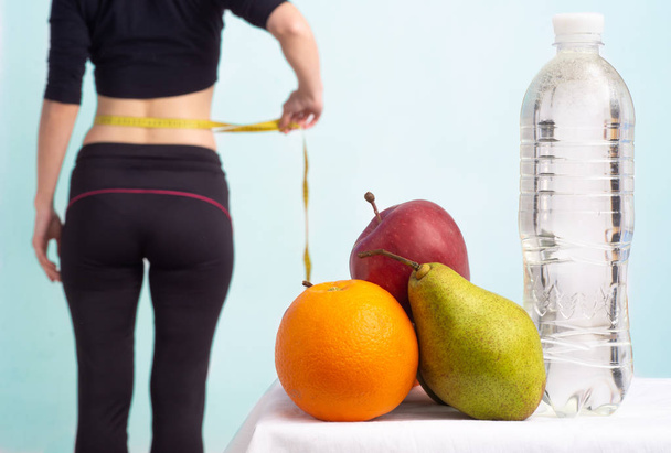 Стройная молодая женщина измеряет свою тонкую талию рулеткой. В фокусе фрукты для потери веса - яблоко, апельсин и груша
.  - Фото, изображение