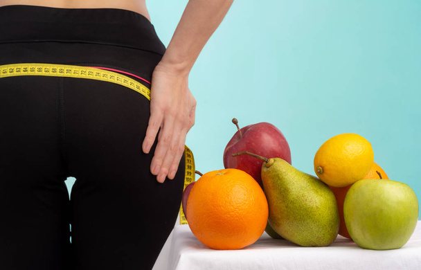 Стройная молодая женщина измеряет свои ягодицы рулеткой. В фокусе фрукты для потери веса - яблоко, апельсин и груша
.  - Фото, изображение