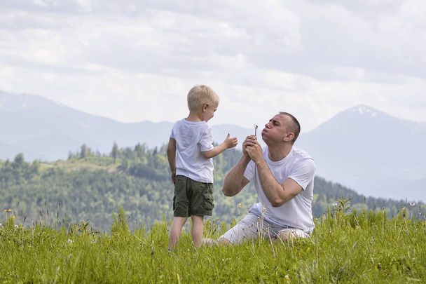 Vader en zoon waait paardebloemen zittend in het gras op een achtergrond van groene bossen, bergen en hemel met wolken. Vriendschap concept. - Foto, afbeelding