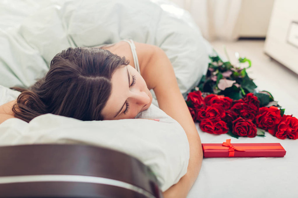 junge Frau schlafend neben Rosenstrauß und Geschenkbox im Bett. Geschenk, das ihr Freund zum Valentinstag hinterlassen hat. Überraschung - Foto, Bild