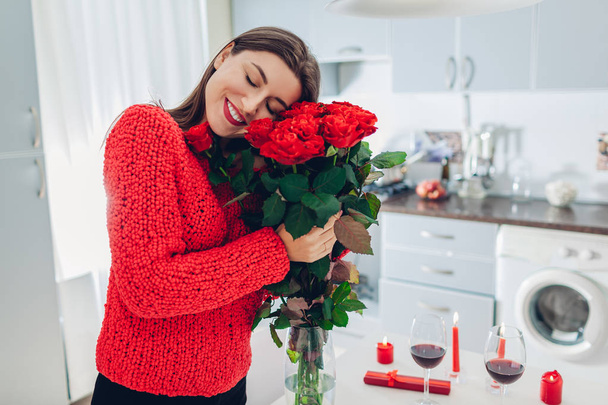 Young woman found bouquet of roses, wine and gift box on kitchen. Happy girl hugging flowers. Valentines day surprise - Фото, зображення