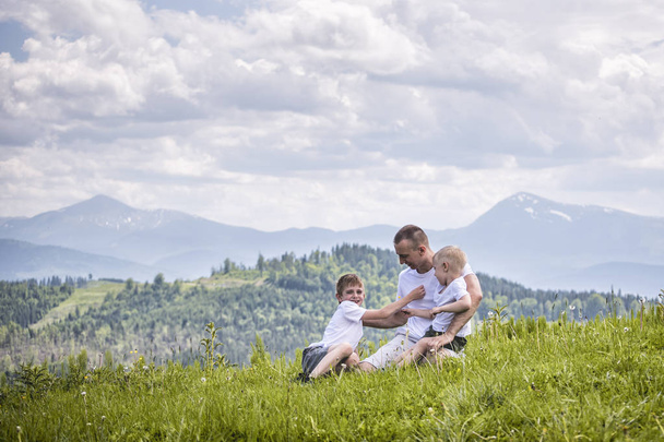 緑の森、山々、空の雲の背景の草の上に座って彼の 2 つの若い息子と父。友情の概念 - 写真・画像