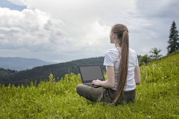 Belle fille avec un ordinateur portable assis sur l'herbe verte sur un fond de montagnes
. - Photo, image