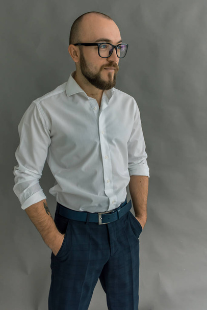 young pretty, stylish man with a beard in glasses in a bright shirt in dark pants on a gray background, emotional and shows movements with his hands - Valokuva, kuva