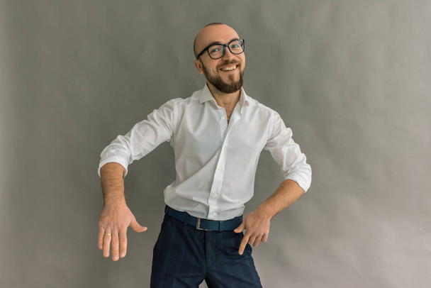 mladý hezký, stylový muž s bradkou v brýlích v světlé tričko v tmavé kalhoty na šedém pozadí, emocionální a ukazuje pohyby rukama - Fotografie, Obrázek