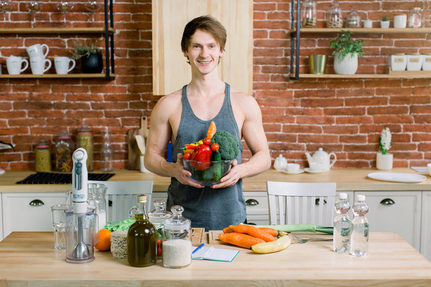 Beau jeune homme souriant en chemise grise tenant un bol avec des légumes frais sur le fond du mur de briques dans la cuisine. Concept de nourriture saine et végétalienne
 - Photo, image