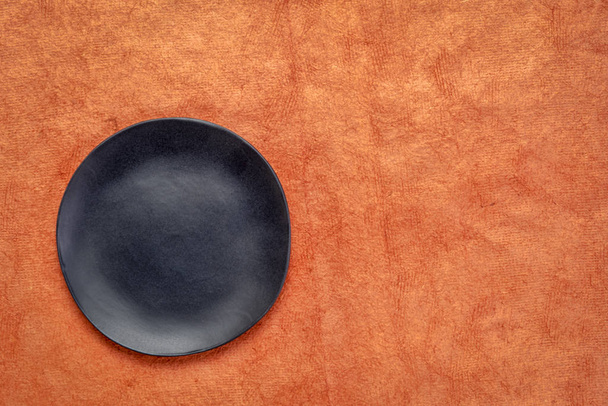 black ceramic plate with an irregular edge on a pumpkin orange handmade Huun Mayan  paper  with a copy space - Foto, afbeelding