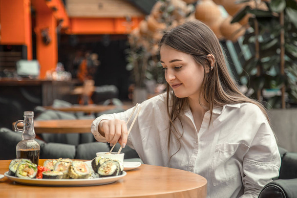 Молодая женщина ест и наслаждается свежими суши в роскошном ресторане. Женщина-клиент держит в руках палочки с едой и ест восточную еду на обед. Концепция питания
. - Фото, изображение
