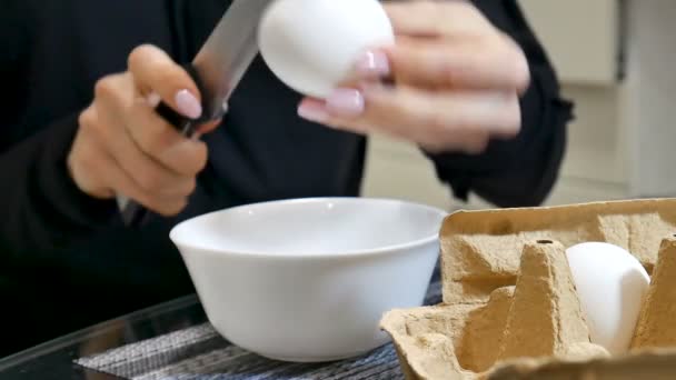 Video z ženských rukou praskliny vajíčko do bílé keramické mísy pomocí nože v kuchyni - Záběry, video