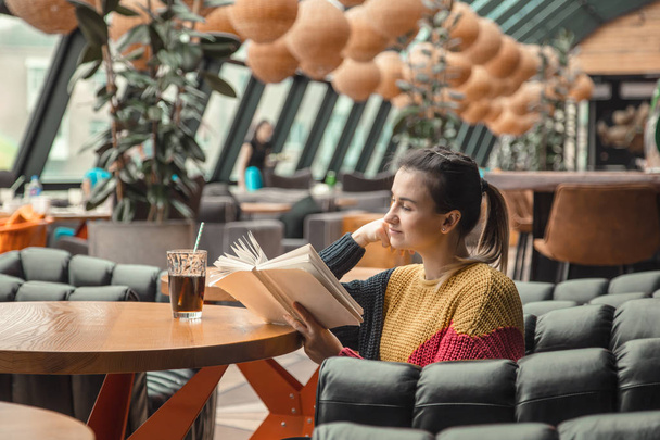 Young beautiful woman in orange sweater reading interesting book in cafe. The concept of reading - Foto, imagen