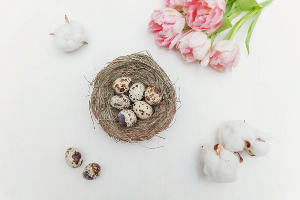 Spring greeting card. Easter eggs in nest cotton and pink fresh tulip flowers bouquet on rustic white wooden background. Easter concept. Flat lay top view copy space. Spring flowers tulips - Фото, зображення
