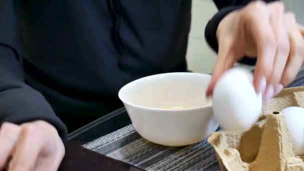 Manos de mujer rompiendo un huevo en un tazón de cerámica blanca en la cocina
 - Metraje, vídeo