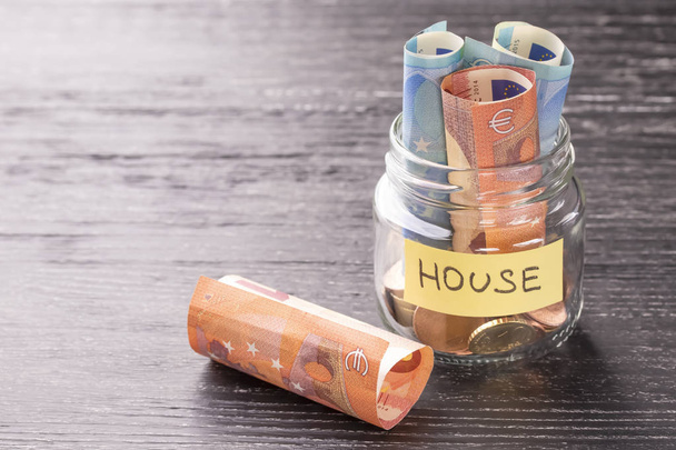 Glasgefäß mit Münzen und Euroscheinen mit dem Wort Haus auf einem schwarzen Holztisch. das Konzept der finanziellen Einsparungen für die Reparatur, den Bau von Haushaltsausgaben. - Foto, Bild