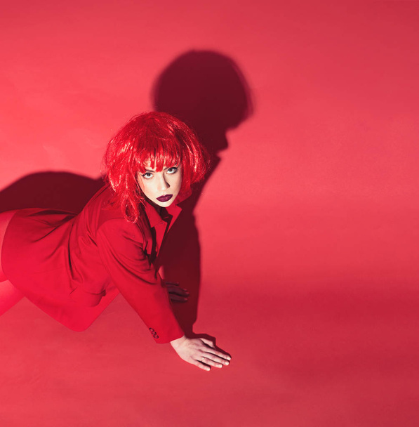 Fashion concept. Lady looking at camera and crawls on floor. Girl on mysterious face in red formal jacket, red background. Woman with makeup and red wig posing in total red outfit, copy space - Foto, Imagem