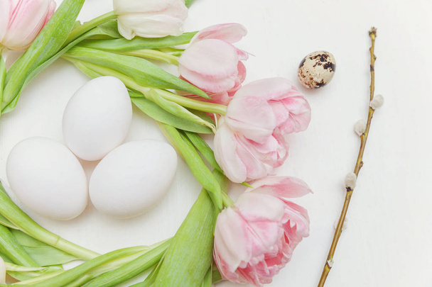 Spring greeting card. Easter eggs in nest and pink fresh tulip flowers bouquet on rustic white wooden background. Easter concept. Flat lay top view copy space. Spring flowers tulips - Foto, Bild
