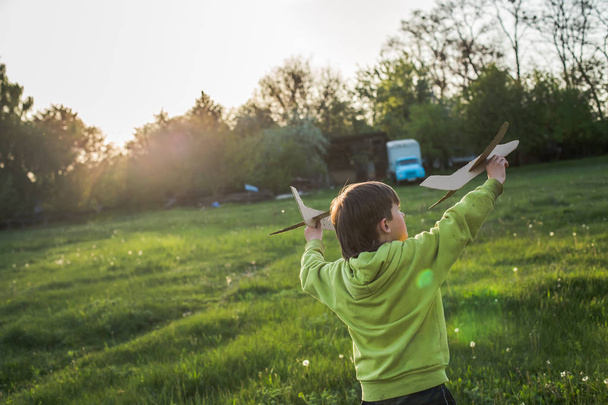 Kind startet Papierflugzeug. Junge in grüner Jacke läuft bei Sonnenuntergang über das Feld. Glückliche Kindheit. Ohne Handy auf der Straße. - Foto, Bild