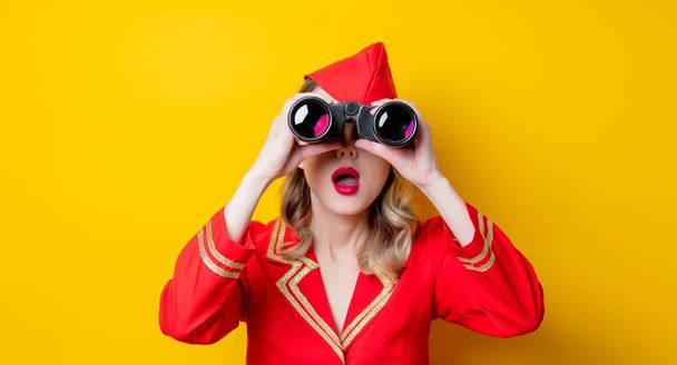 Retrato de una encantadora azafata vintage con uniforme rojo y prismáticos. Aislado sobre fondo amarillo
. - Foto, Imagen