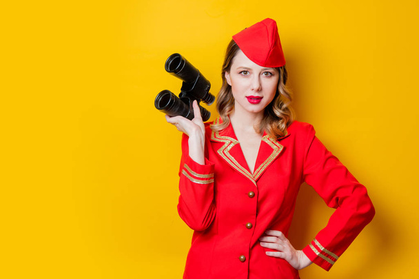 Portrait of charming vintage stewardess wearing in red uniform with binoculars. Isolated on yellow background. - Фото, изображение