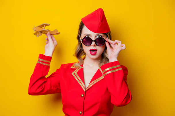 Portrait of charming vintage stewardess wearing in red uniform and sunglasses with airplane. Isolated on yellow background. - Foto, afbeelding