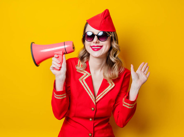Πορτρέτο της πανέμορφο vintage αεροσυνοδός φορώντας κόκκινο στολή με τηλεβόα. Απομονωμένα σε κίτρινο φόντο. - Φωτογραφία, εικόνα