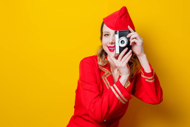 Portrait of charming vintage stewardess wearing in red uniform with photo camera. Isolated on yellow background. - Photo, Image