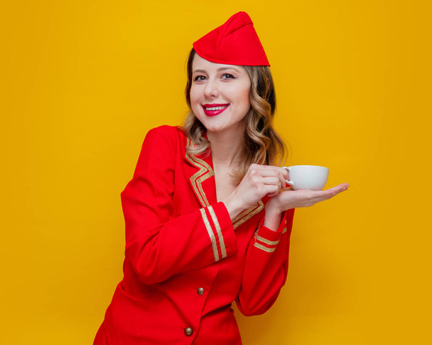 Kırmızı bardak kahve veya çay ile üniforma giyen büyüleyici vintage hostes portresi. Sarı arka plan üzerinde izole. - Fotoğraf, Görsel