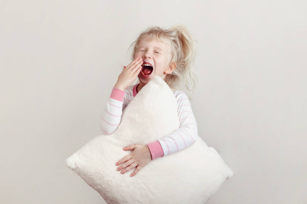 Healthy sleep concept. Cute blonde Caucasian girl child in pink pajamas with closed eyes yawning covering wide open mouth with palm. Sleepy kid with messy hair holding white soft pillow - Photo, Image