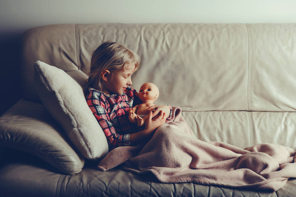 Child playing with toy. Cute Caucasian girl holding cuddling with baby doll. Kid lying in bed at evening night time indoors. Toned with film filters style. Happy childhood lifestyle. - Photo, Image